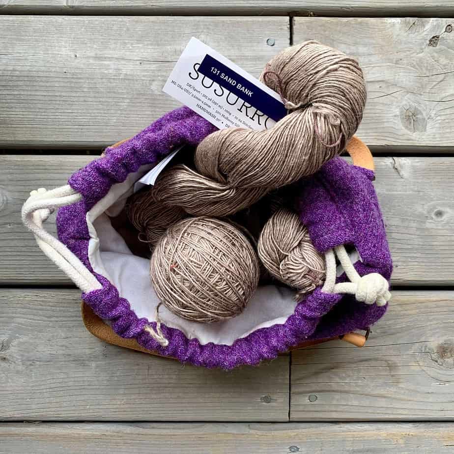 Ball of beige yarn and two skeins of the same yarn peek out of the top of a purple drawstring bag. The bag is on a wooden surface and the photograph is looking directly down into the bag. The lining and drqstring of the bag are white. The text on the blue label on the skein of yarn reads sand bank. The white label below reads Susurro though the letters are slightly obscured by the label above.