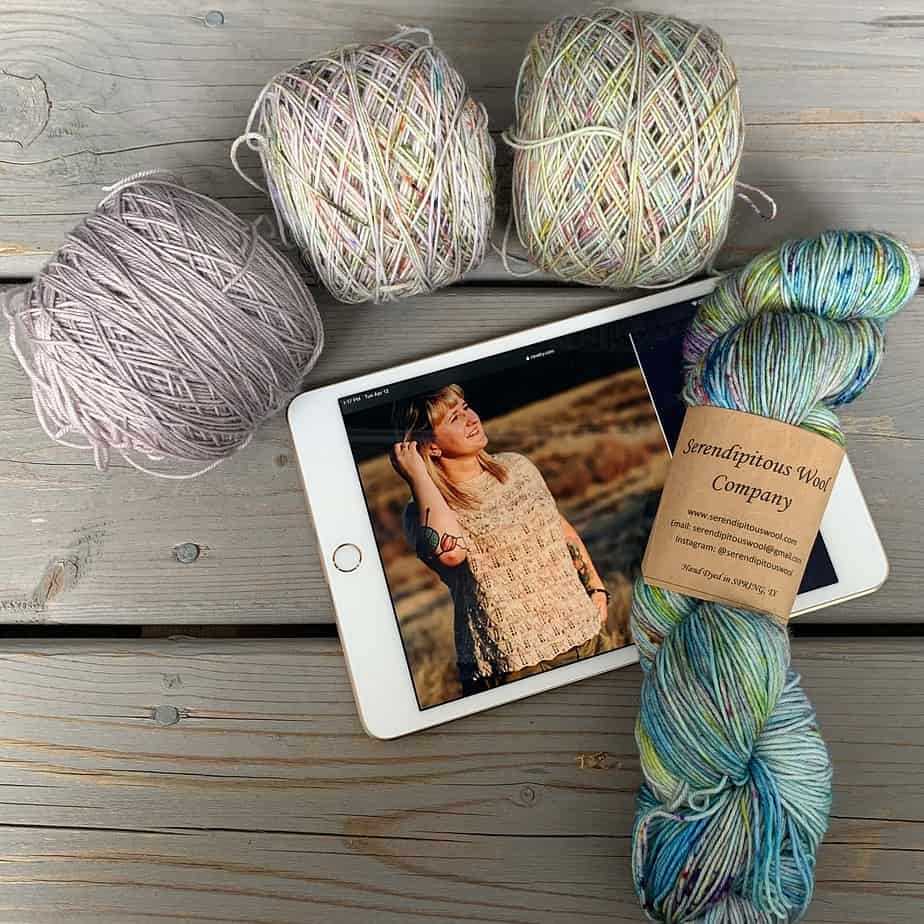 An ipad lies on a wooden surface with a photo of a white woman tucking her dark blonde hair behind her right ear.She is wearing a lace patterned short sleeve knitted top in a creamy beige colour.