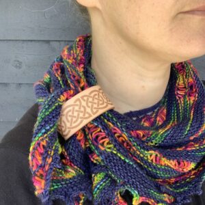 A close cropped photo of a white woman wearing a navy and rainbow coloured knitted shawl. She is looking to her left and only her lips and chin are visible. Her shawl is held in place by a pale honey coloured leather cuff with a celtic knot design etched onto it.