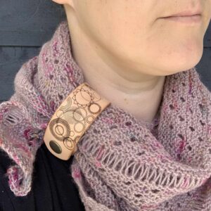 A close cropped photo of a white woman wearing a dusty pink coloured knitted shawl. She is looking to her left and only her lips and chin are visible. Her shawl is held in place by a pale honey coloured leather cuff with a collection of many circular shapes etched onto it.