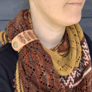 A close cropped photo of a white woman wearing a gold, white and rust red coloured knitted shawl. She is looking to her left and only her lips and chin are visible. Her shawl is held in place by a pale honey coloured leather cuff with the outline of four feathers etched onto it.