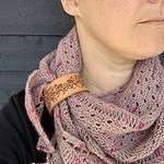 A close cropped photo of a white person wearing a dusty pink coloured knitted shawl. Only their neck is visible. The shawl is held in place by a pale honey coloured leather cuff with a flower and leaf design etched onto it.