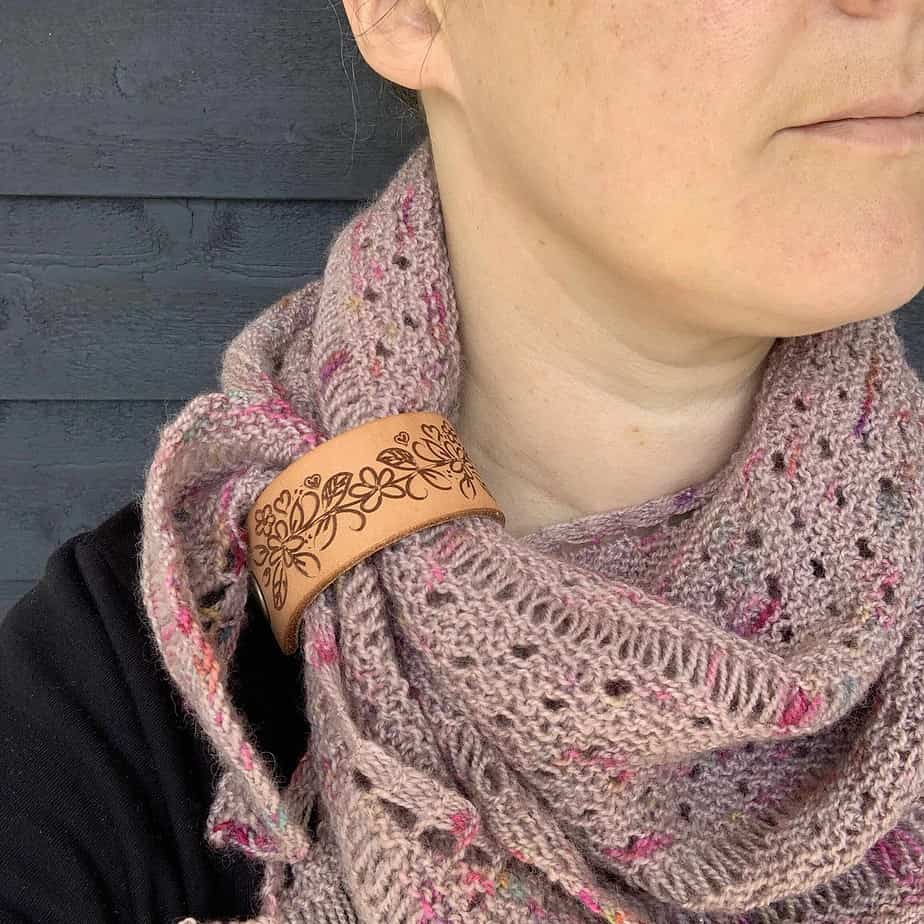 A close cropped photo of a white person wearing a dusty pink coloured knitted shawl. Only their neck is visible. The shawl is held in place by a pale honey coloured leather cuff with a flower and leaf design etched onto it.
