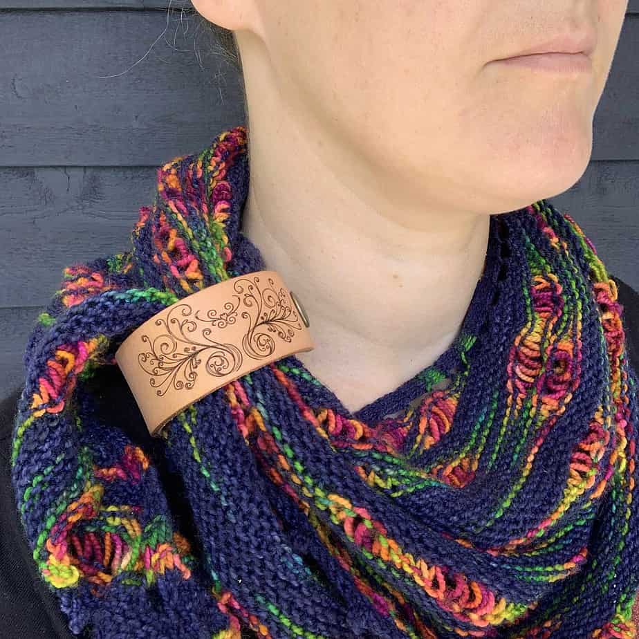 A close cropped photo of a white woman wearing a navy and rainbow coloured knitted shawl. She is looking to her left and only her lips and chin are visible. Her shawl is held in place by a pale honey coloured leather cuff showing a heart shape with many flourishes.