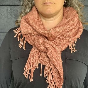 A white woman wearing a dark long-sleeved top stands in front of a dark wooden surface. Her eyes are not visible in the photograph. She is wearing a pink knitted shawl knotted around her neck. The fringed edges of the shawl are visible.