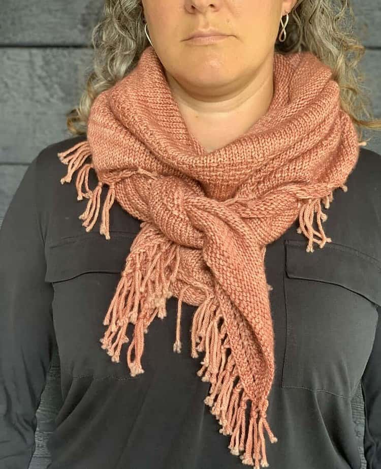 A white woman wearing a dark long-sleeved top stands in front of a dark wooden surface. Her eyes are not visible in the photograph. She is wearing a pink knitted shawl knotted around her neck. The fringed edges of the shawl are visible.