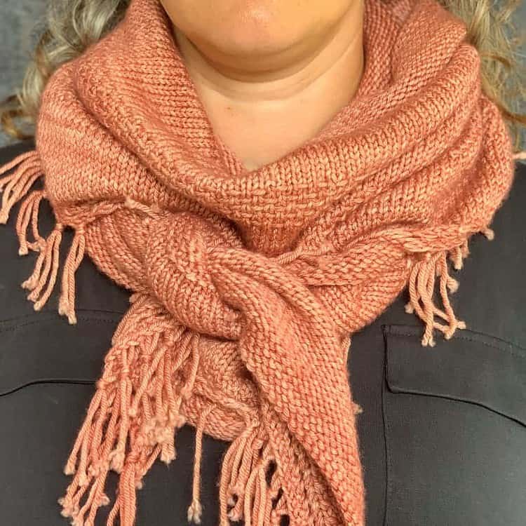 A white woman wearing a dark top has a pink knitted shawl knotted around her neck. The fringed edges of the shawl are visible.