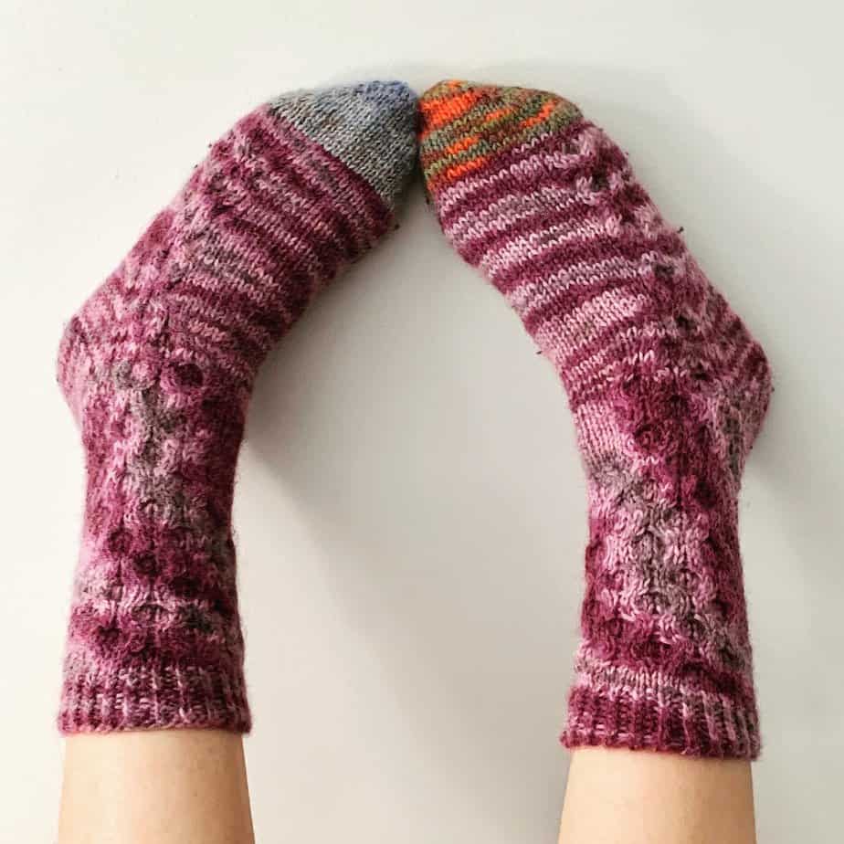 A closeup of the feet of a white person wearing pink and purple coloured knitted socks. Their feet are against a whit wall with their toes pointing towards each other.