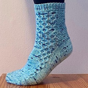 A closeup view of a person wearing black trousers and standing on tiptoes on a wooden surface. Only one foot is visible and turned to the left of the photograph to show the side of the foot. They are wearing a pale blue knitted sock with dark blue speckles. The sock has a honeycomb cable pattern with begins at the top cuff and tapers off to a single cable running to the smallest toe.