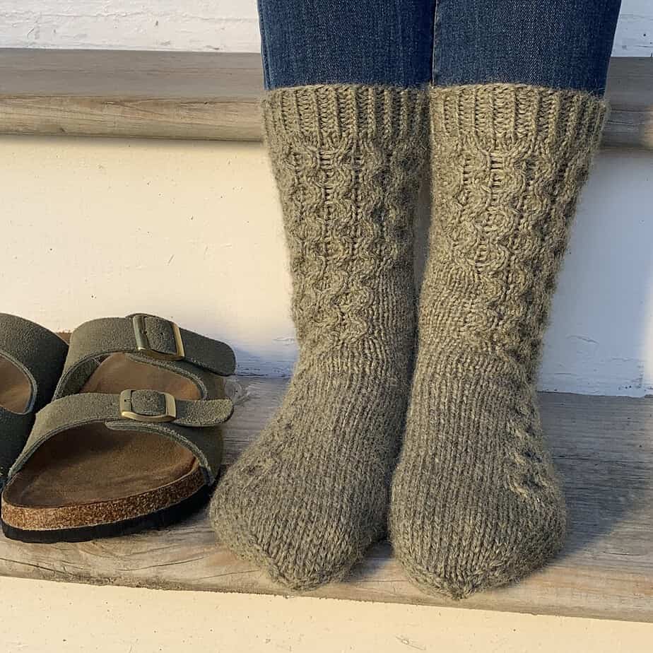 A closeup image of a person's feet as they are sitting on a wooden step. There is a pair of khaki green sandals on the step to the left of their feet. They are wearing olive green knitted socks with a cable pattern which begins at the top of the sock and tapers down to the smallest toe on each foot.