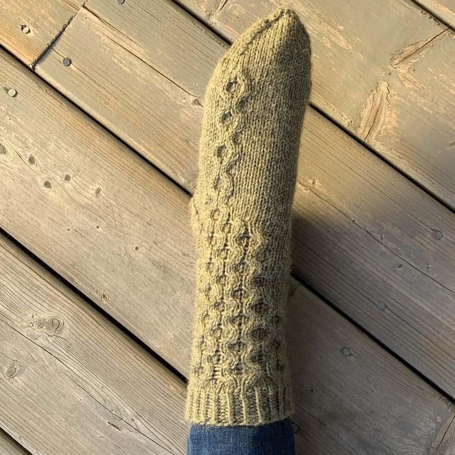 A closeup view of a left foot stretched out with pointed toes on a wooden surface. The foot is wearing an olive green knitted sock with honeycomb cables running down the outside toward the smallest toe.
