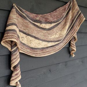 A knitted shawl hjangs on a dark painted wooden wall. The shawl is knitted in charcoal, pink and peach coloured stripes with some wedge shaped lace sections.