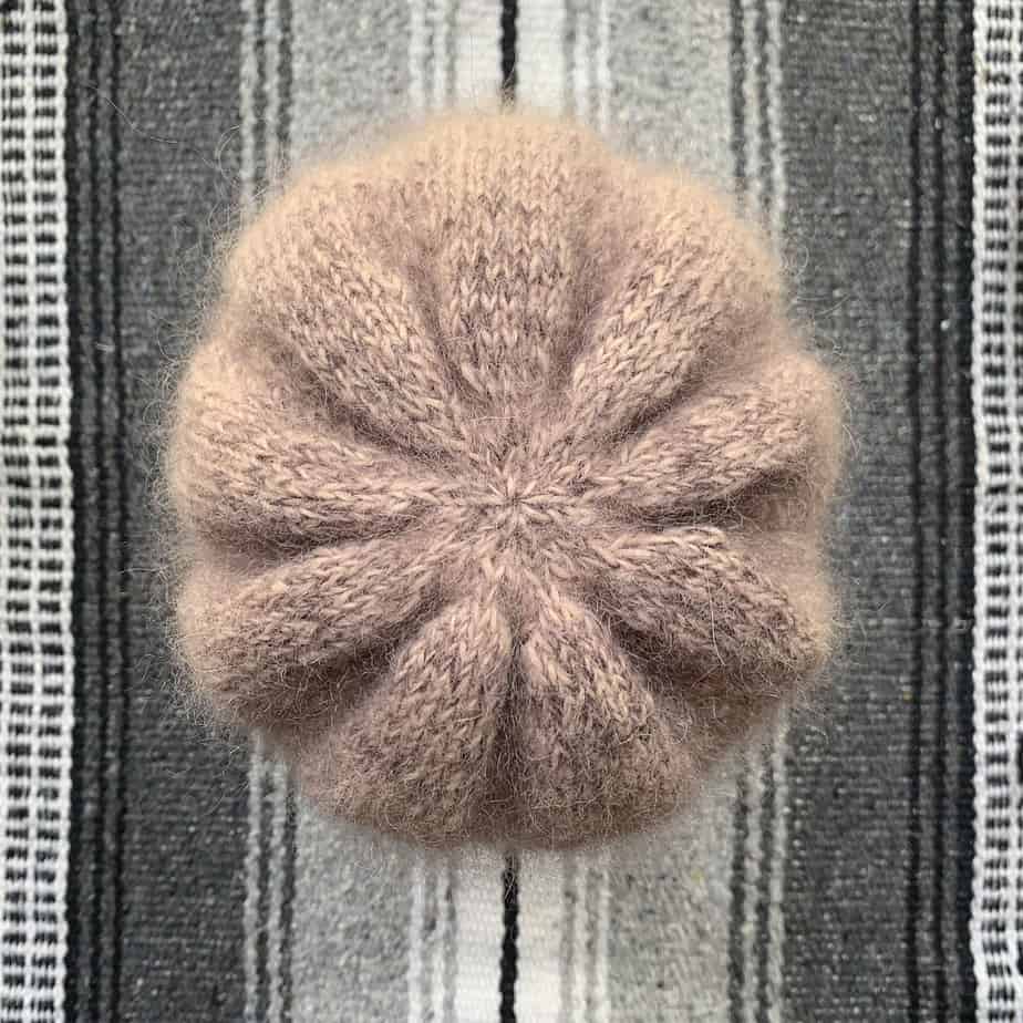 The camera is looking down on the crown of a dusky pink knitted hat. The blanket underneath the hat is striped in many shades of black, white and grey. The hat has a gathered crown which is shaped like a flower with nine petals. The fluffy halo of the fabric is clearly visible.