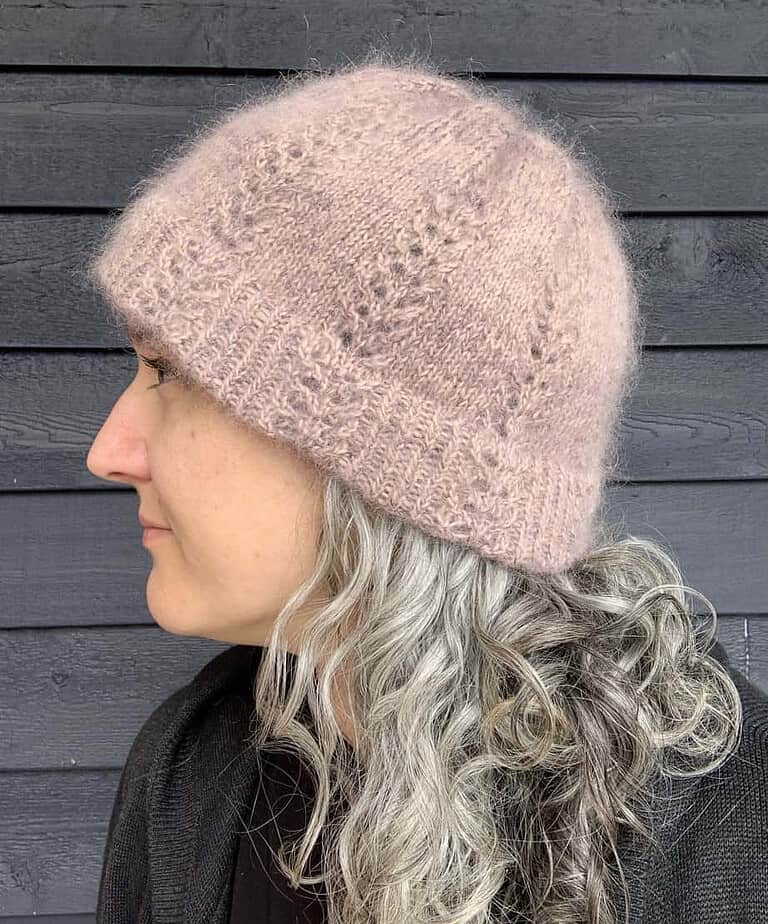 A white woman with silver and brown hair stands in front of a dark painted wooden surface looking to her right, only her profile is visible. She is wearing a black top and a fluffy, dusky pink knitted hat which has a linear lace pattern reaching towards the gathered crown.