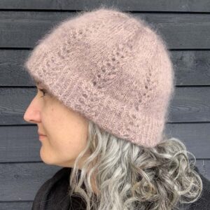 A white woman with silver and brown hair stands in front of a dark painted wooden surface looking to her right, only her profile is visible. She is wearing a black top and a fluffy, dusky pink knitted hat which has a linear lace pattern reaching towards the gathered crown.