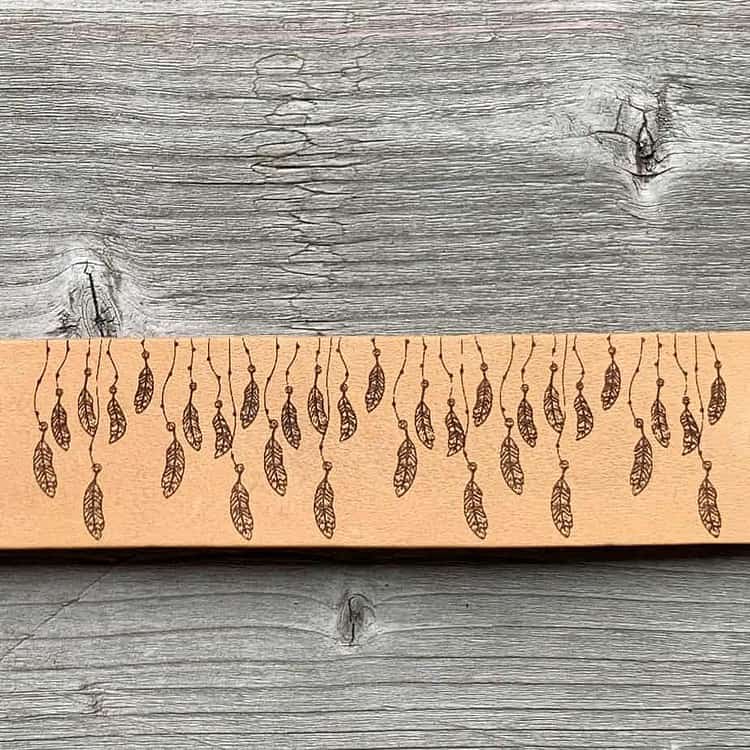 A close-up view of a natural undyed leather cuff on a grey wooden surface. There is a design etched onto the cuff showing thirty-three tiny feathers hanging from strings.