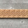 A close-up view of a natural undyed leather cuff on a grey wooden surface. There is a design etched onto the cuff showing an elongated celtic band of linked knots.