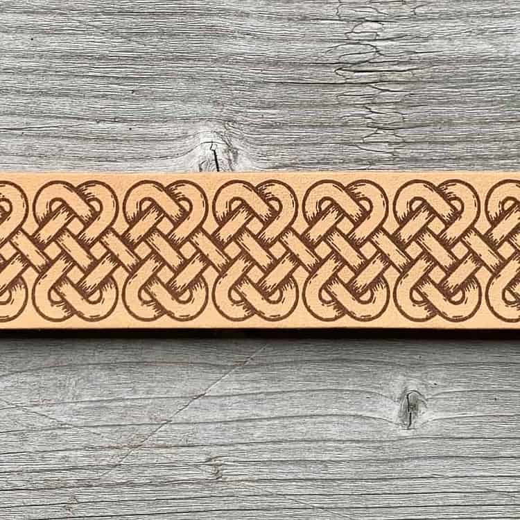 A close-up view of a natural undyed leather cuff on a grey wooden surface. There is a design etched onto the cuff showing an elongated celtic band of linked knots.