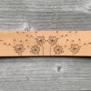 A close-up view of a natural undyed leather cuff on a grey wooden surface. There is a design etched onto the cuff showing six dandelion clocks with their seeds flying in a breeze.
