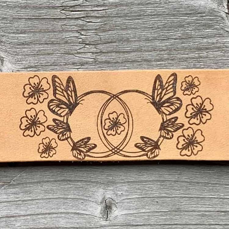 A close-up view of a natural undyed leather cuff on a grey wooden surface. There is a design etched onto the cuff showing six butterflies gathered around two linked circles and surrounded by flowers.