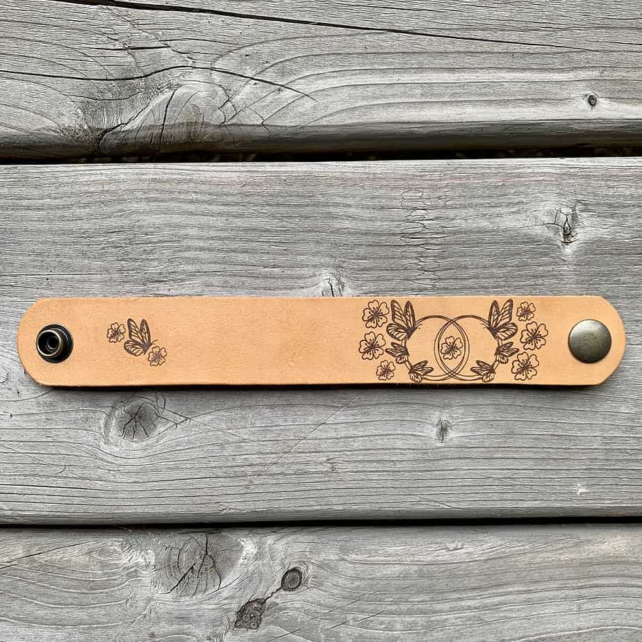 A natural undyed leather cuff with a snap closure on either end lies flat on a grey wooden surface. There is a design etched onto the cuff showing six butterflies gathered around two linked circles and surrounded by flowers.