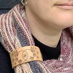 A close cropped photo of a white woman wearing a pink, peach and charcoal coloured knitted shawl. She is looking to her left and only her lips and chin are visible. Her shawl is held in place by a pale honey coloured leather cuff with an engraved design showing butterlies flying around two linked circles with a flower in the centre.