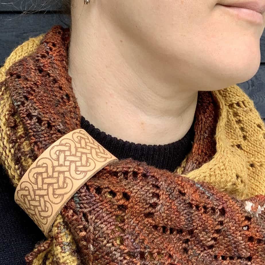 A close cropped photo of a white woman wearing a warm coloured knitted shawl in autumnal shades of rust and yellow. She is looking to her left and only her lips and chin are visible. Her shawl is held in place by a pale honey coloured leather cuff with an engraved design of a long celtic knot.