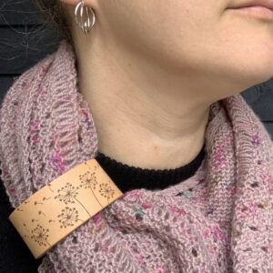 A close cropped photo of a white woman wearing a dusty pink coloured knitted shawl. She is looking to her left and only her lips and chin are visible. Her shawl is held in place by a pale honey coloured leather cuff with a design featuring dandelion clocks blowing in a breeze engraved onto it.