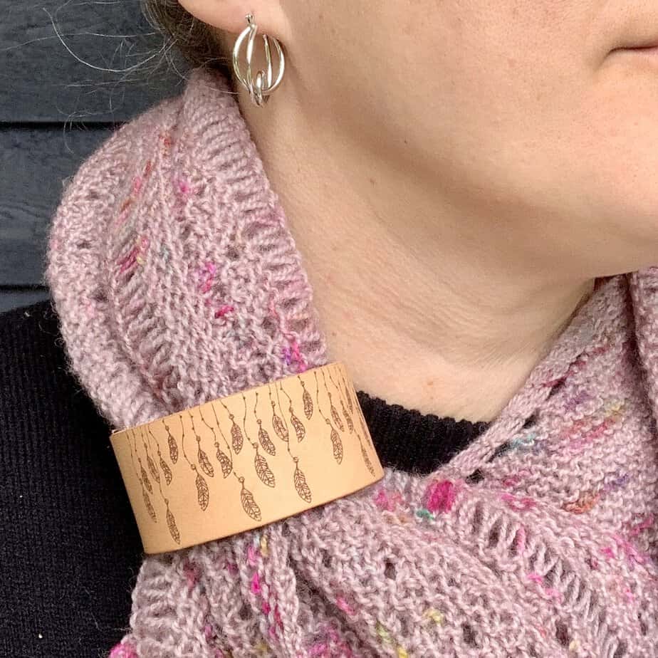 A close cropped photo of a white woman wearing a dusty pink coloured knitted shawl. She is looking to her left and only her lips and chin are visible. Her shawl is held in place by a pale honey coloured leather cuff with a design featuring feathers hanging from strings engraved onto it.