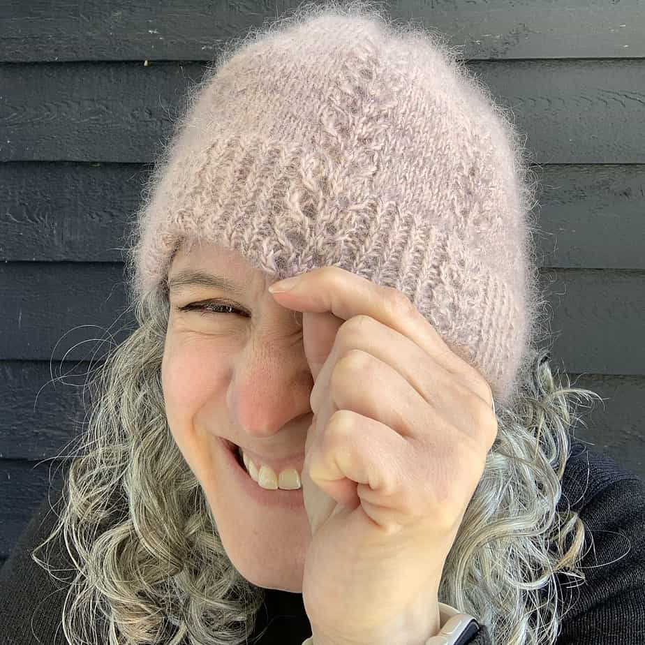 A white woman with silver and black hair grins at the camera. She is wearing a dusky pink fluffy hat and is pulling it down over her left eye with her left hand. She is very proud of the hat she has knitted.