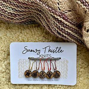 A white rectangular piece of card sits on a pale yellow sheepskin surface. There is a knitted shawl in shades of dusky pink and beige at the top of the image. The large text on the white card reads Snowy Thistle Knits. There is a grey strand of wool strung through two holes in the card. Five colourful lightbulb shaped pins are threaded onto the grey wool. The pins are gold, rose gold, orange, bright red and deep red. Each pin is holding a silver ring attached to a small round wooden charm. The wooden charms have single images of celtic knots laser engraved onto the surface. Tiny text printed on the card below the charms reads stitch markers marqueurs de point.