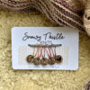 A white rectangular piece of card sits on a pale yellow sheepskin surface. There is a knitted shawl in shades of dusky pink and beige at the top of the image. The large text on the white card reads Snowy Thistle Knits. There is a grey strand of wool strung through two holes in the card. Five colourful lightbulb shaped pins are threaded onto the grey wool. The pins are gold, orange, rose gold, bright red and deep red. Each pin is holding a silver ring attached to a small round wooden charm. The wooden charms have single images of a tea cup, a sheep, a sun, a snowflake and a heart laser engraved onto the surface. Tiny text printed on the card below the charms reads stitch markers marqueurs de point.