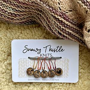 A white rectangular piece of card sits on a pale yellow sheepskin surface. There is a knitted shawl in shades of dusky pink and beige at the top of the image. The large text on the white card reads Snowy Thistle Knits. There is a grey strand of wool strung through two holes in the card. Five colourful lightbulb shaped pins are threaded onto the grey wool. The pins are gold, orange, rose gold, bright red and deep red. Each pin is holding a silver ring attached to a small round wooden charm. The wooden charms have single images of a tea cup, a sheep, a sun, a snowflake and a heart laser engraved onto the surface. Tiny text printed on the card below the charms reads stitch markers marqueurs de point.
