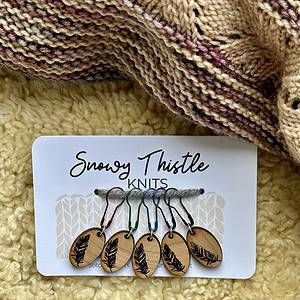 A white rectangular piece of card sits on a pale yellow sheepskin surface. There is a knitted shawl in shades of dusky pink and beige at the top of the image. The large text on the white card reads Snowy Thistle Knits. There is a grey strand of wool strung through two holes in the card. Five colourful lightbulb shaped pins are threaded onto the grey wool. The pins are brown, green, blue, purple and black. Each pin is holding a silver ring attached to a small oval wooden charm. The wooden charms have single images of feathers laser engraved onto the surface. Tiny text printed on the card below the charms reads stitch markers marqueurs de point and is barely visible between the markers.