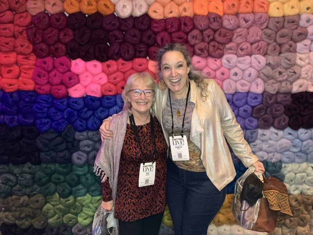 Two women wearing huge smiles are standing in front of a wall covered in rainbow-coloured fluffy balls of yarn. They seem to be very excited. They are dressed for a party and wearing lanyards with large white labels showing Knitting Live in large print. They are carrying shiny tote bags stuffed full of yarn, magazines and other unknown goodies
