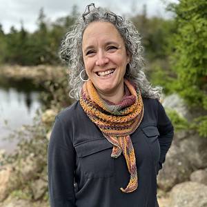 I white woman with silvery hair stands in front of a tree-lined lake. The sky is grey but she is smiling because she is wearing a pretty striped, knitted shawl. The orange, green and claret colours of the shawl compliment the colours in the background.