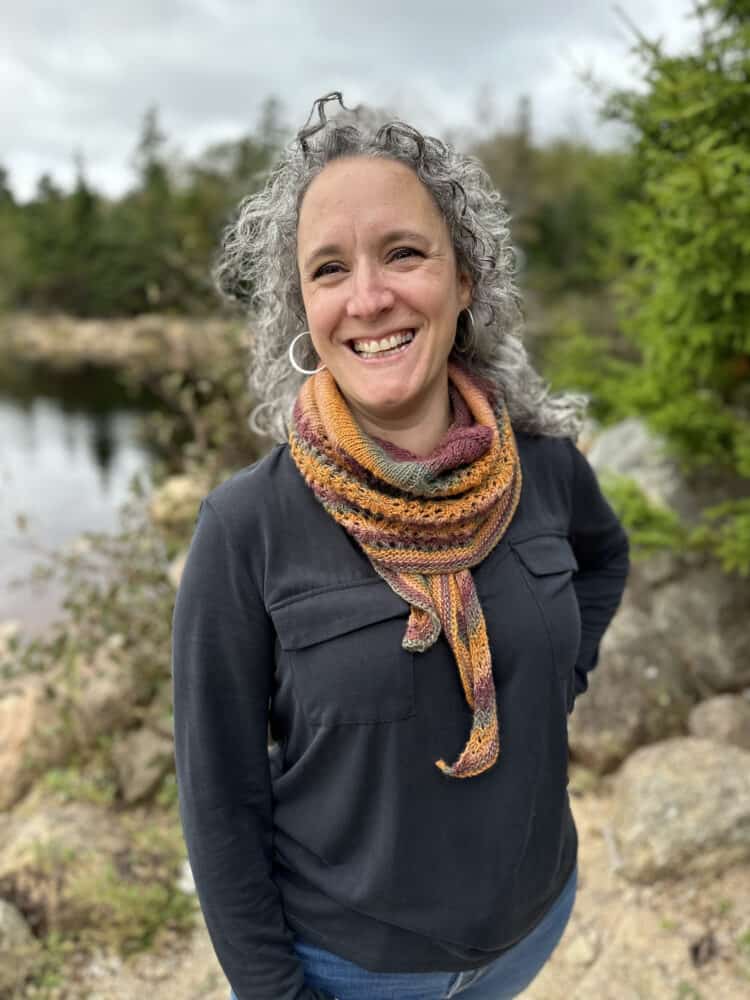 I white woman with silvery hair stands in front of a tree-lined lake. The sky is grey but she is smiling because she is wearing a pretty striped, knitted shawl. The orange, green and claret colours of the shawl compliment the colours in the background.