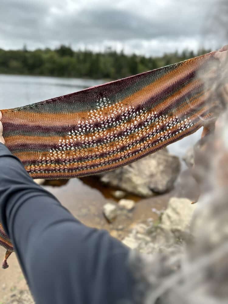 Two outstretched arms hold up a small, stripey, knitted shawl so that the light from the grey sky and lake in the background can shine through the triangular lace section of the shawl and illuminate the eyelets. The stripes of burnt orange, fir green and claret compliment the background of the image.