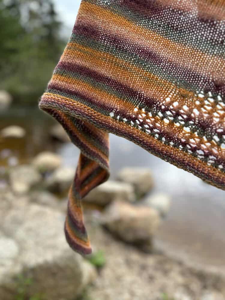 A close up image of the curly end of a knitted shawl suspended in the air over the edge of a muddy tree-lined lake.The shawl is striped in shades of burnt orange, fir green and claret which compliment the background of the image. There is a small section of lace visible from the middle of the shawl.
