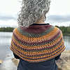 A white woman with curly silvery hair stands with her back to the camera. She is looking out upon a tree-lined lake and an expansive grey sky. Her hair is swept onto her left shoulder to show the knitted shawl she wears draped across her shoulders. The stripes of burnt orange, fir green and claret are a bright contrast to the background of the image. The shawl has a triangular wedge of lace stitches across the middle of the knitter's back.