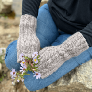 A close up image of a person's hands as they are resting on their knees. The person is wearing blue jeans and a black long-sleeved shirt. The photo is focussed on the knitted fingerless mittens worn by the subject. The mittens are pale grey. The fabric is ribbed, with occasional cable crosses where the straight lines of the ribbing cross to go in a different direction. The person is also holding a tiny bunch of purple daisy-like flowers.