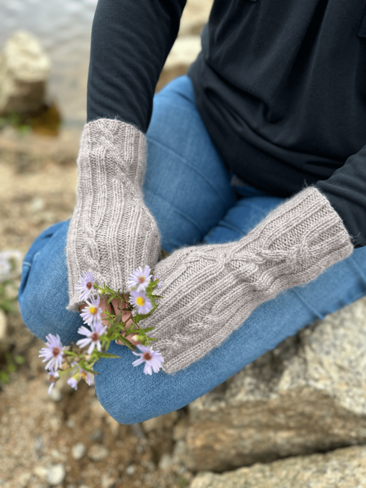 A close up image of a person's hands as they are resting on their knees. The person is wearing blue jeans and a black long-sleeved shirt. The photo is focussed on the knitted fingerless mittens worn by the subject. The mittens are pale grey. The fabric is ribbed, with occasional cable crosses where the straight lines of the ribbing cross to go in a different direction. The person is also holding a tiny bunch of purple daisy-like flowers.