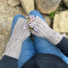 A close up image, taken from above, looking down onto a person's hands as they are resting on their knees. The person is wearing blue jeans and a black long-sleeved shirt. The photo is focussed on the pale grey, knitted, fingerless mittens worn by the subject. The fabric is ribbed, with occasional cable crosses where the straight lines of the ribbing cross to go in a different direction. The person is also holding a tiny bunch of purple daisy-like flowers. The background of the photo shows many rocks in different sizes and shapes.