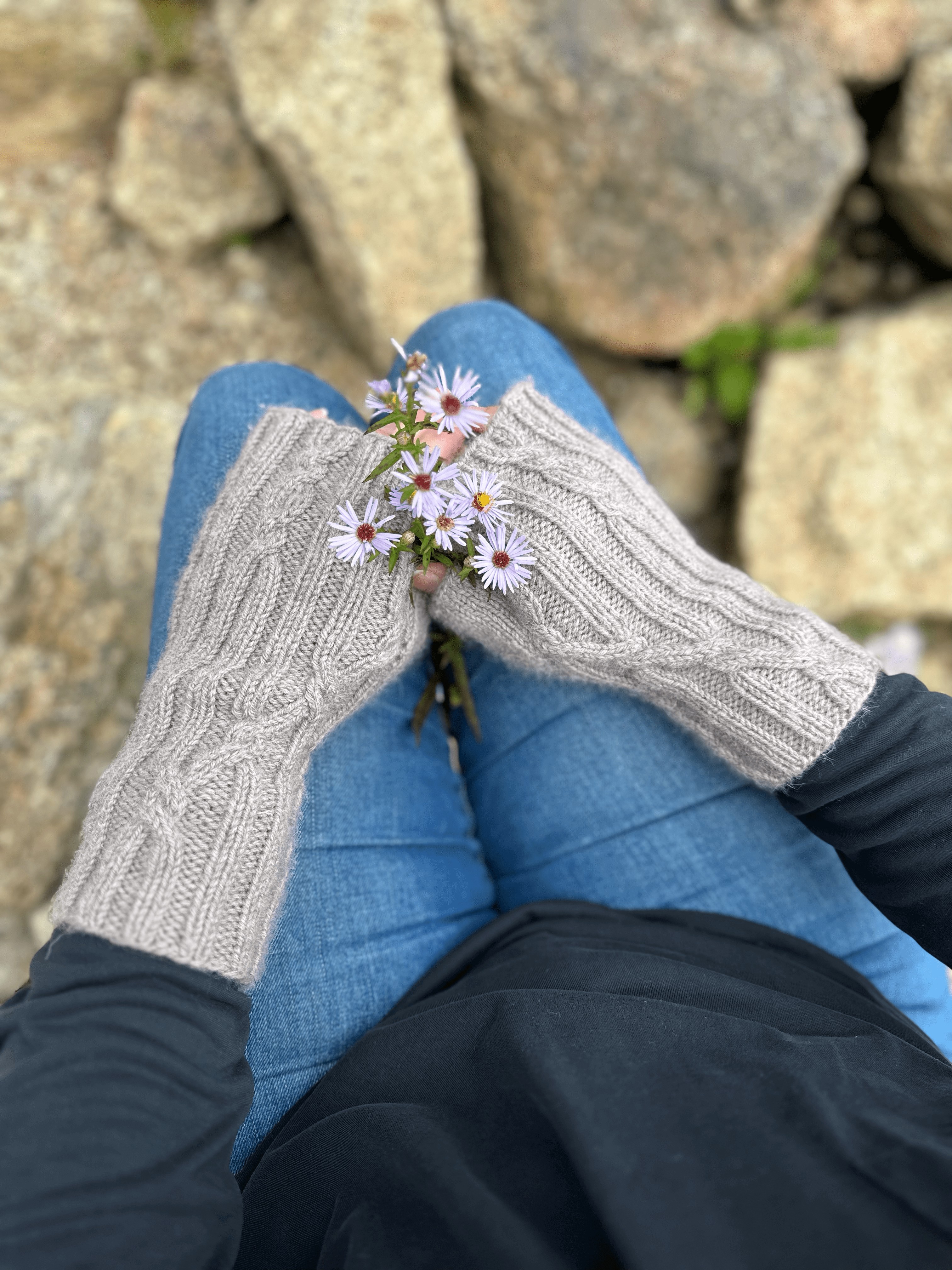 A close up image, taken from above, looking down onto a person's hands as they are resting on their knees. The person is wearing blue jeans and a black long-sleeved shirt. The photo is focussed on the pale grey, knitted, fingerless mittens worn by the subject. The fabric is ribbed, with occasional cable crosses where the straight lines of the ribbing cross to go in a different direction. The person is also holding a tiny bunch of purple daisy-like flowers. The background of the photo shows many rocks in different sizes and shapes.