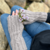 A close up image, taken from above and behind the subject's left shoulder, looking down onto a person's hands as they are resting on their knees. The person is wearing blue jeans and a black long-sleeved shirt. The photo is focussed on the pale grey, knitted, fingerless mittens worn by the subject. The fabric is ribbed, with occasional cable crosses where the straight lines of the ribbing cross to go in a different direction. The person is also holding a tiny bunch of purple daisy-like flowers. The background of the photo shows many rocks in different sizes and shapes.