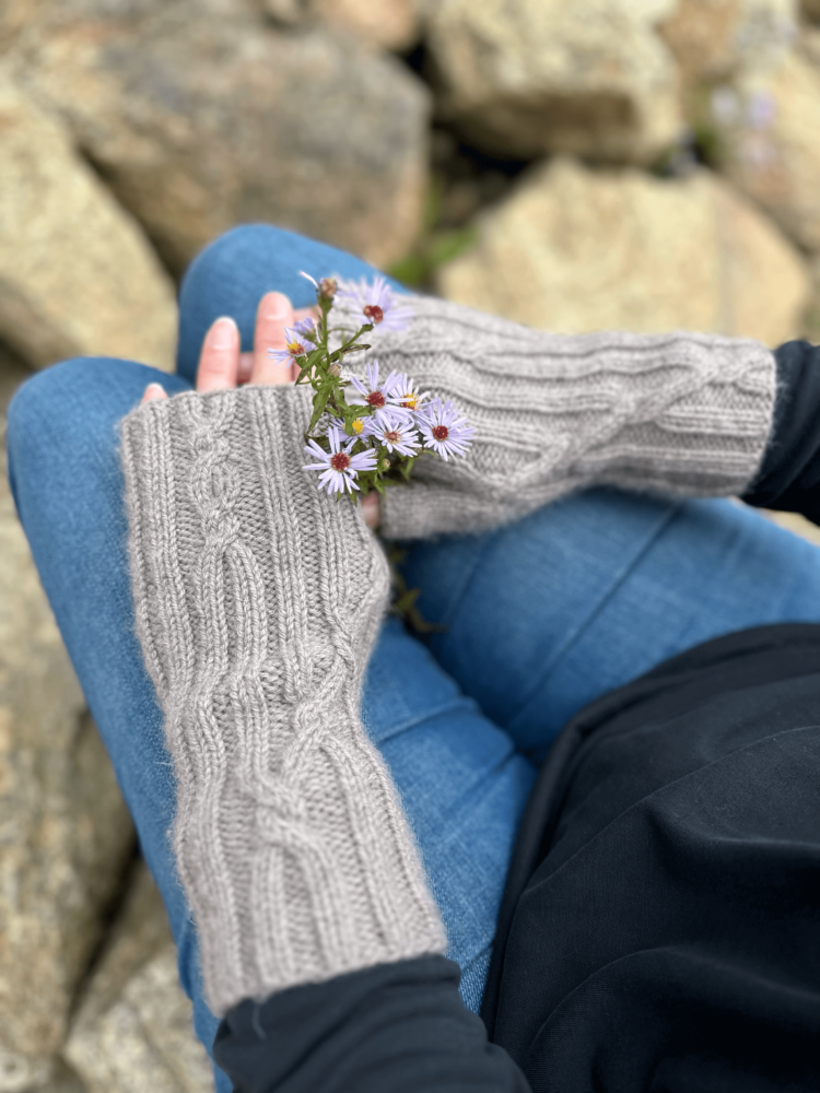 A close up image, taken from above and behind the subject's left shoulder, looking down onto a person's hands as they are resting on their knees. The person is wearing blue jeans and a black long-sleeved shirt. The photo is focussed on the pale grey, knitted, fingerless mittens worn by the subject. The fabric is ribbed, with occasional cable crosses where the straight lines of the ribbing cross to go in a different direction. The person is also holding a tiny bunch of purple daisy-like flowers. The background of the photo shows many rocks in different sizes and shapes.