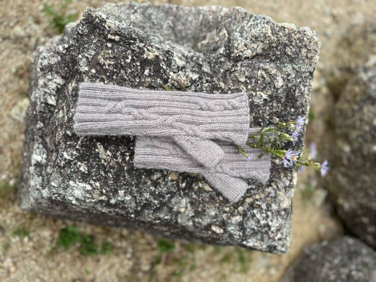 A pair of pale grey knitted mittens are lying on a speckled grey stone. The mittens are fingerless and the fabric is ribbed with a few