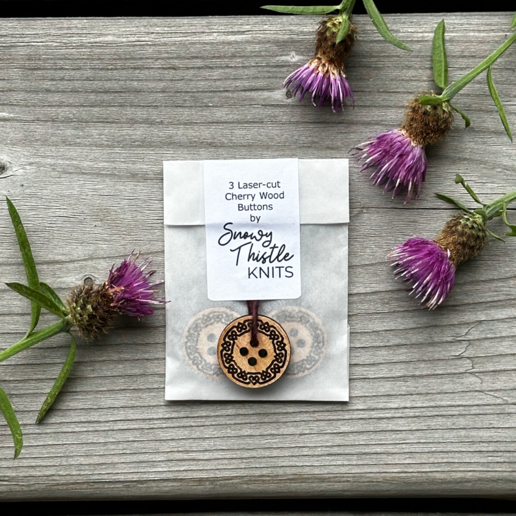 Image shows a small, white, semi-translucent paper package lying on a grey wooden surface surrounded by 4 thistle heads. There are two wooden buttons visible through the paper package. A white label is affixed to the opening of the package with text which reads 3 laser-cut cherry wood buttons by Snowy Thistle Knits. The label holds in place a length of dark brown yarn which is threaded through one hole of a single cherry wood button on the outside of the package. There is an interlocking celtic hearts pattern around the edge of the circular button.