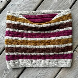 A knitted cowl lies on a pale grey wooden surface. The cowl has a basketweave texture and is knitted in stripes of pale grey, ochre, brown and fuchsia. The middle of the top edge is folded down to show it has a fuchsia pink lining.