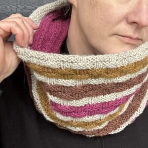 A white person wears a black sweater with a knitted cowl around their neck. It is striped in pale grey, ochre, brown and fucshia. There is a basketweave pattern on the outside of the cowl. The inside lining of the cowl is fuchsia pink. They are holding the cowl slightly to the left of the photo with their hand to allow the lining to be seen.